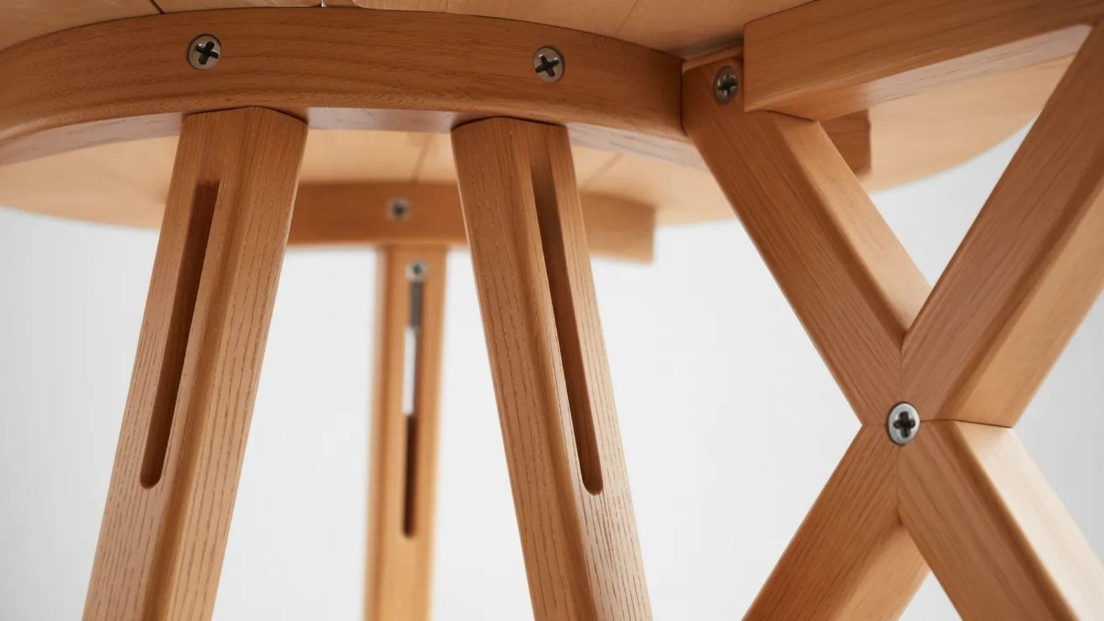 Close-up view of a wooden table's underside, showcasing intersecting slotted legs with screws. The light wood tones create a modern, minimalist feel.