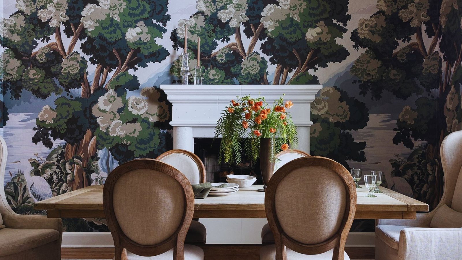 Dining room with a wooden table and four upholstered chairs, set against a forest-themed mural. A flower arrangement on the table adds a fresh touch.