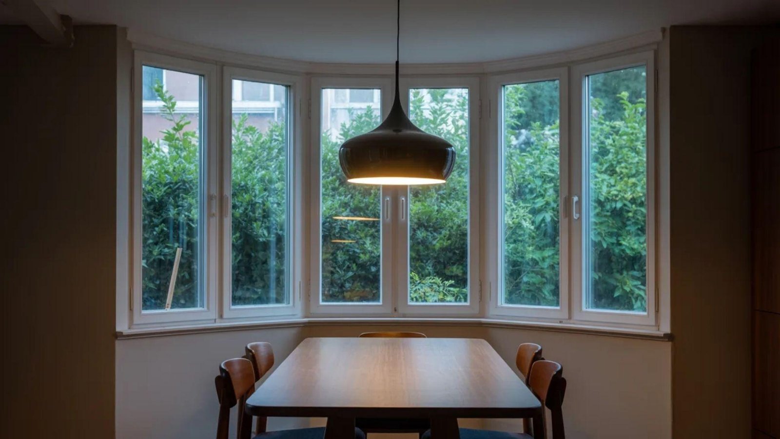 A cozy dining area with a wooden table and four chairs is lit by a pendant lamp. Large bay windows reveal lush green foliage outside, creating a tranquil atmosphere.