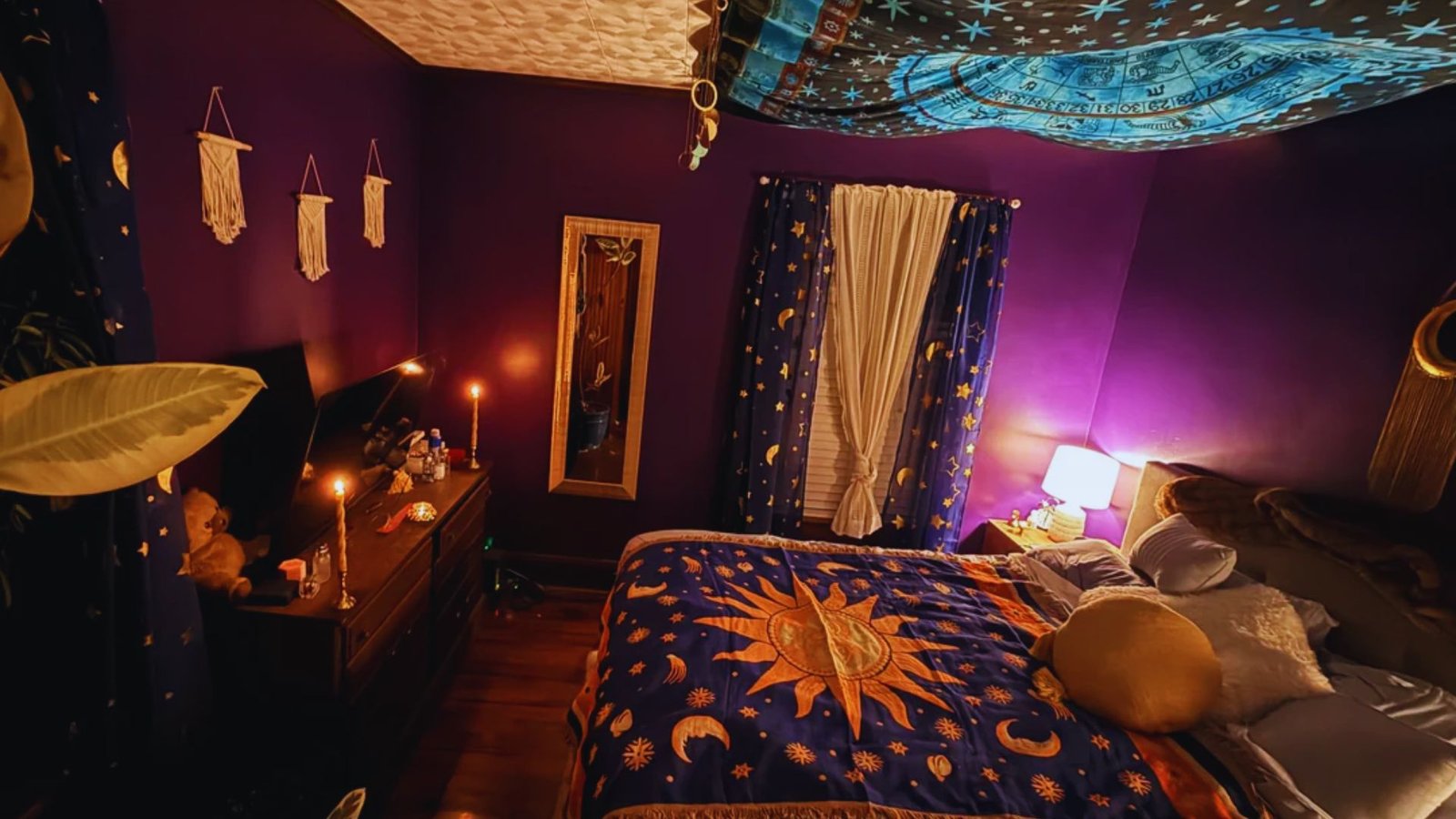 A cozy bedroom with deep purple walls and celestial decor. The bedspread features a sun and moon design. Warm lighting creates a serene, mystical ambiance.