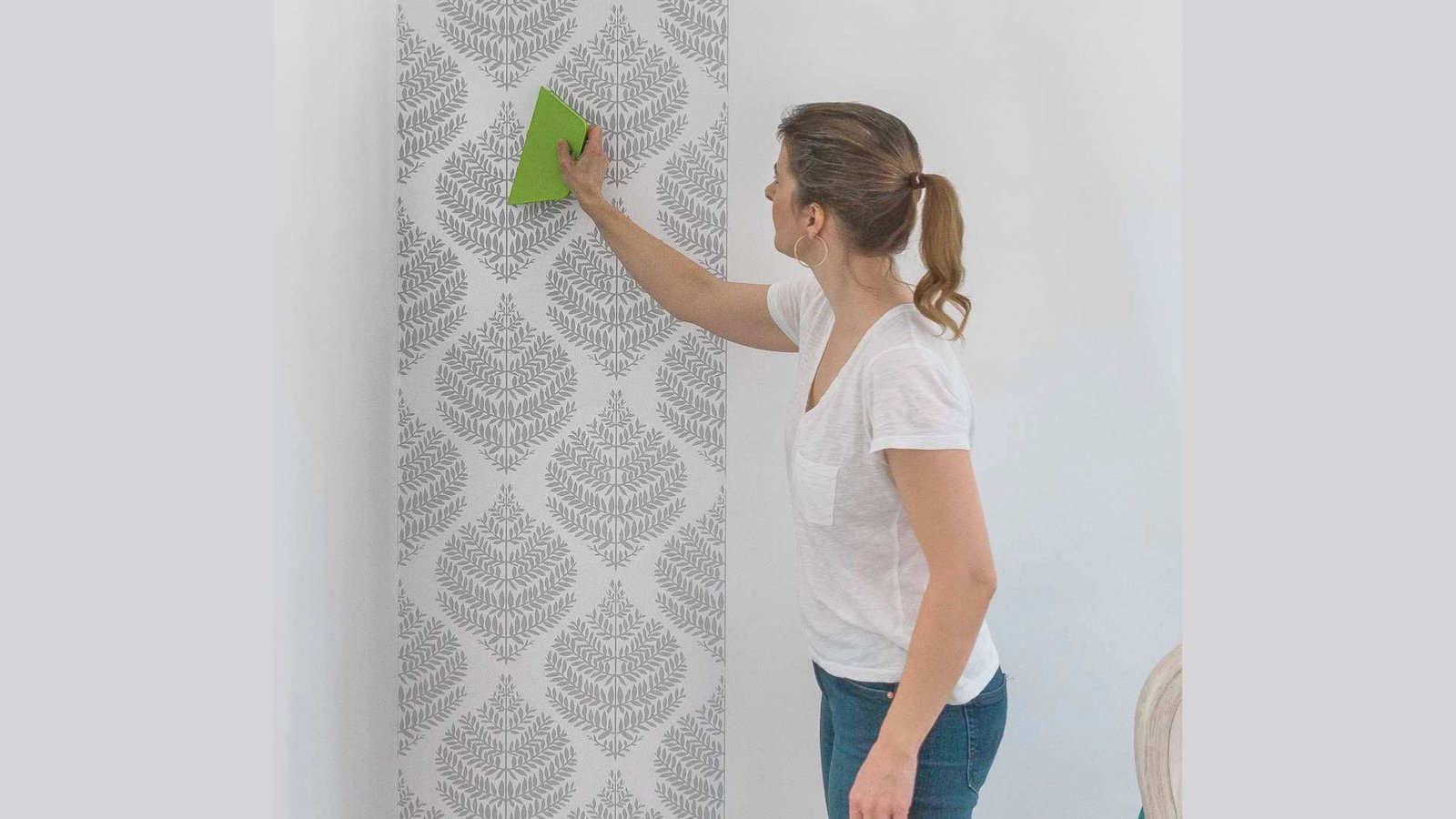 A woman smooths wallpaper with a green tool on a white wall featuring a gray damask pattern. She's focused, wearing a white shirt and blue jeans.