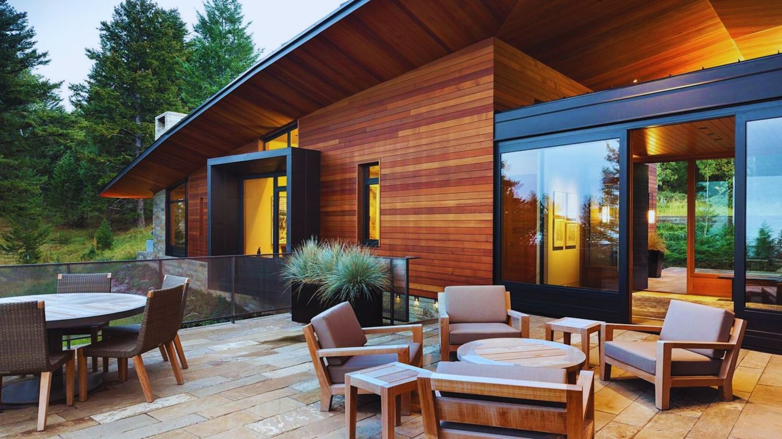 Modern wooden house with large windows on a patio surrounded by trees. Outdoor seating, warm lighting, and a peaceful, inviting atmosphere.