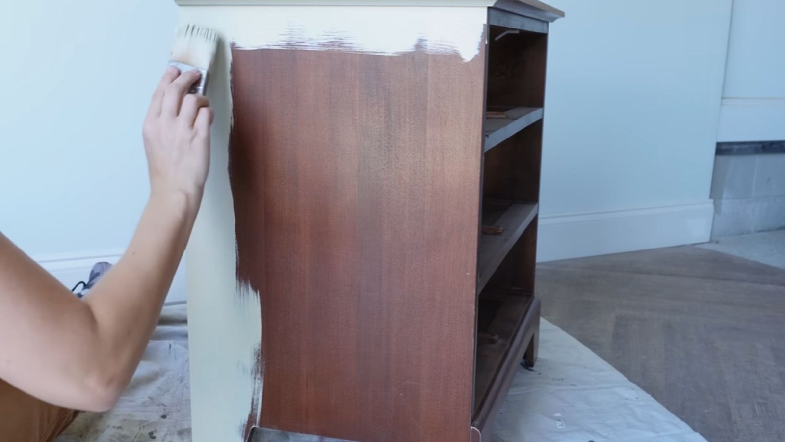 A hand paints the side of a wooden dresser with a white brush. The dresser is partially covered in white paint against a light blue wall, suggesting a DIY project.