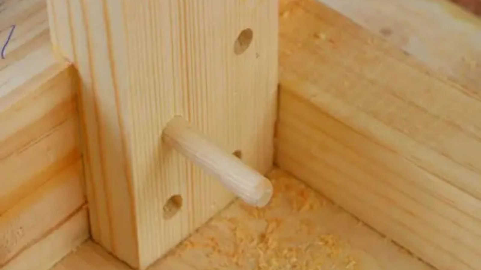 A wooden dowel protrudes through aligned holes in intersecting planks, surrounded by sawdust. This setup conveys a practical woodworking scene.