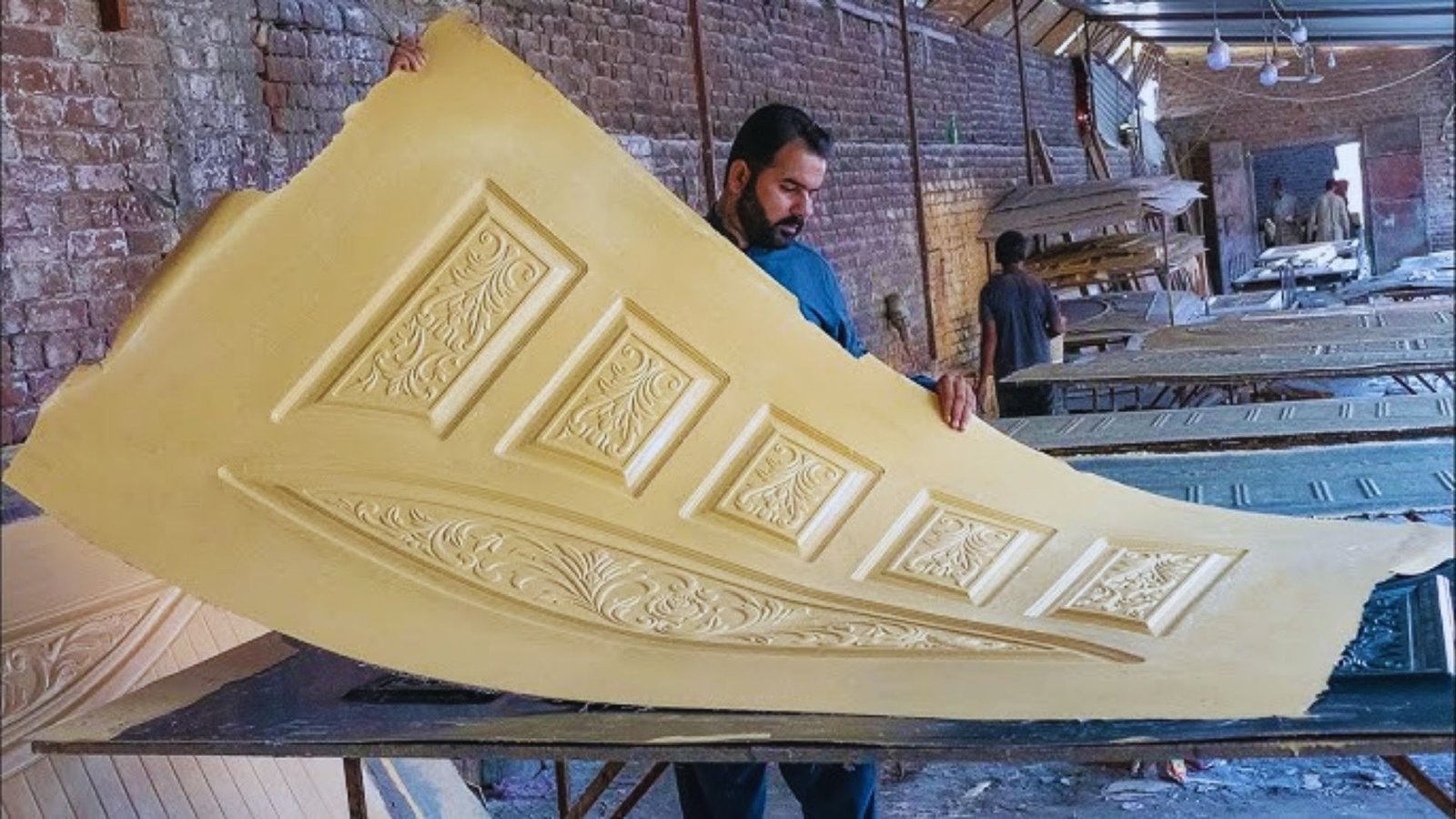 A man in a workshop holds a large, curved decorative ceiling panel with intricate designs. The brick walls and wooden tables create an artisanal atmosphere.