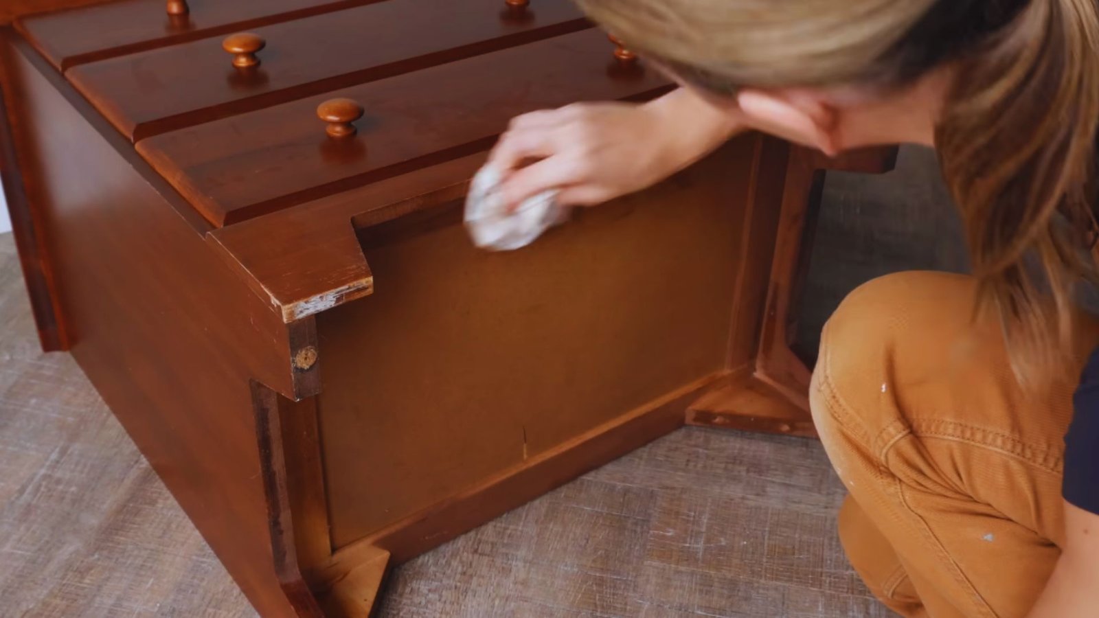 A person cleans a wooden chest with a cloth, focusing on a worn corner. The scene conveys care and restoration, set on a wooden floor.