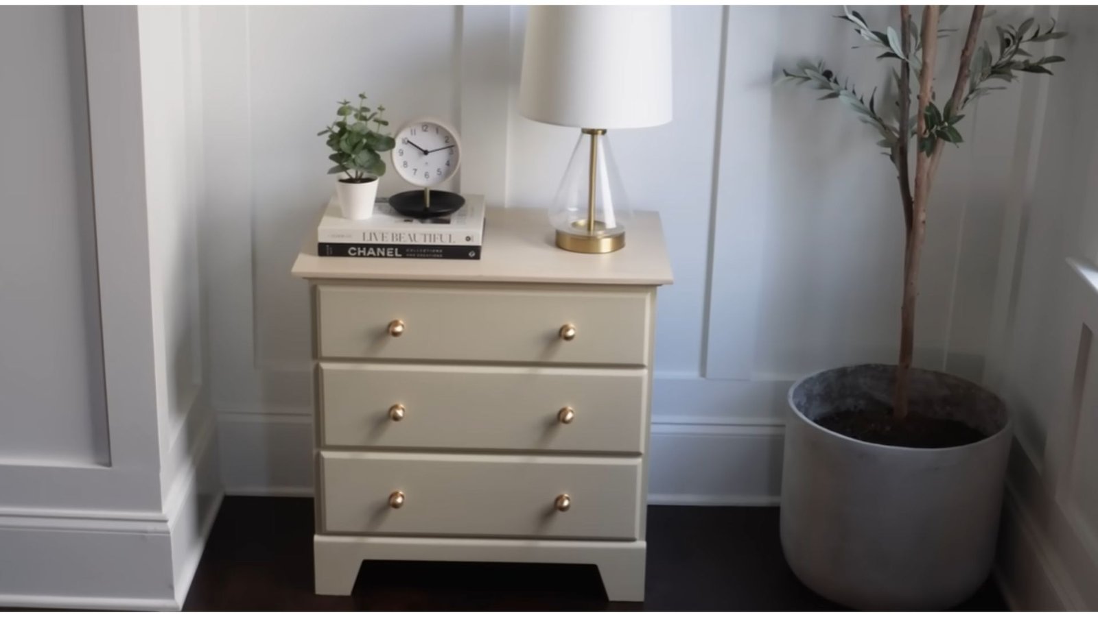 A beige nightstand with gold knobs features a lamp, clock, plant, and books. Nearby is a tall potted plant, set against white paneled walls.