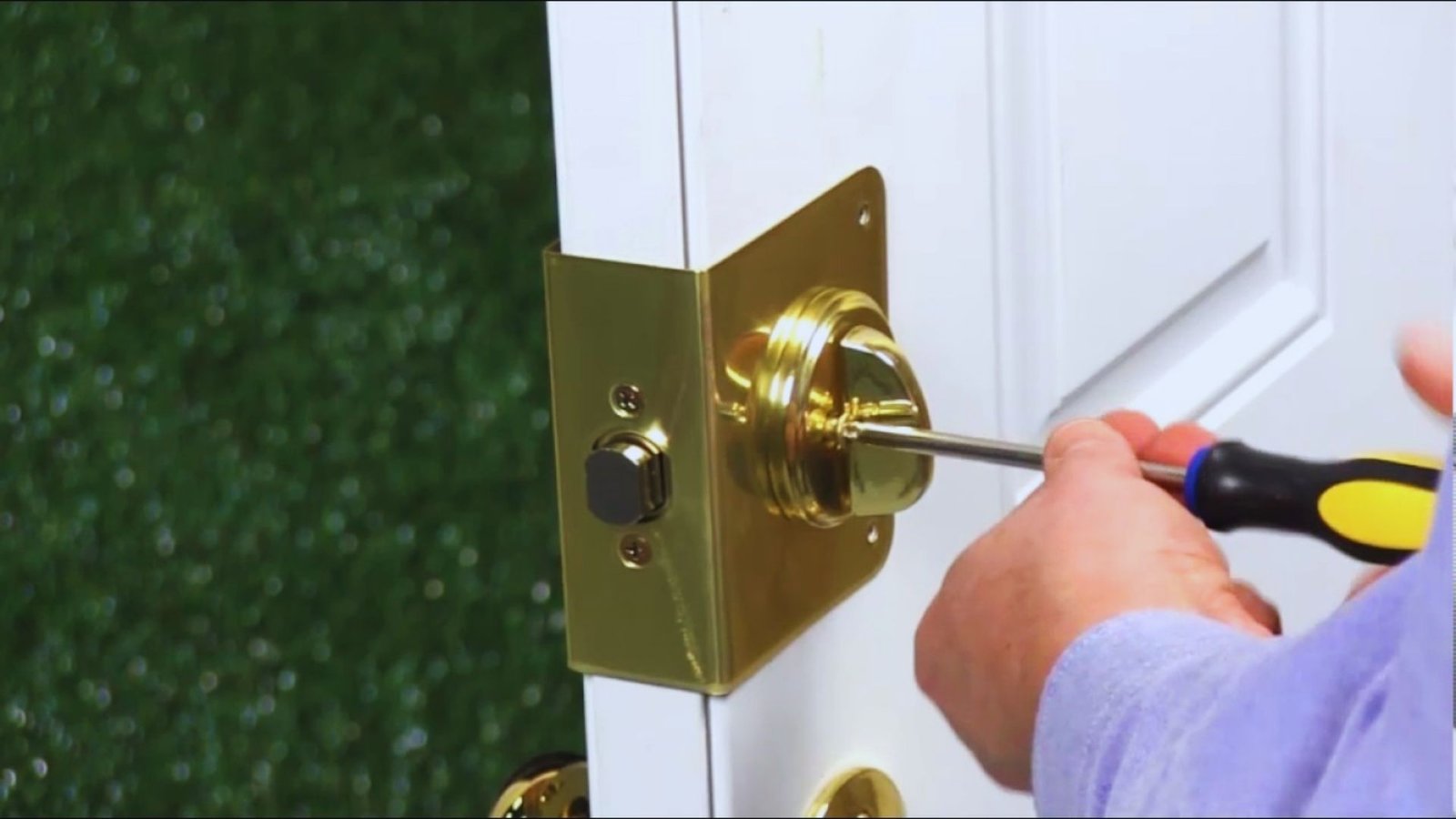 A person in a blue sleeve uses a screwdriver to install a shiny brass deadbolt lock on a white door, with green grass visible in the background.