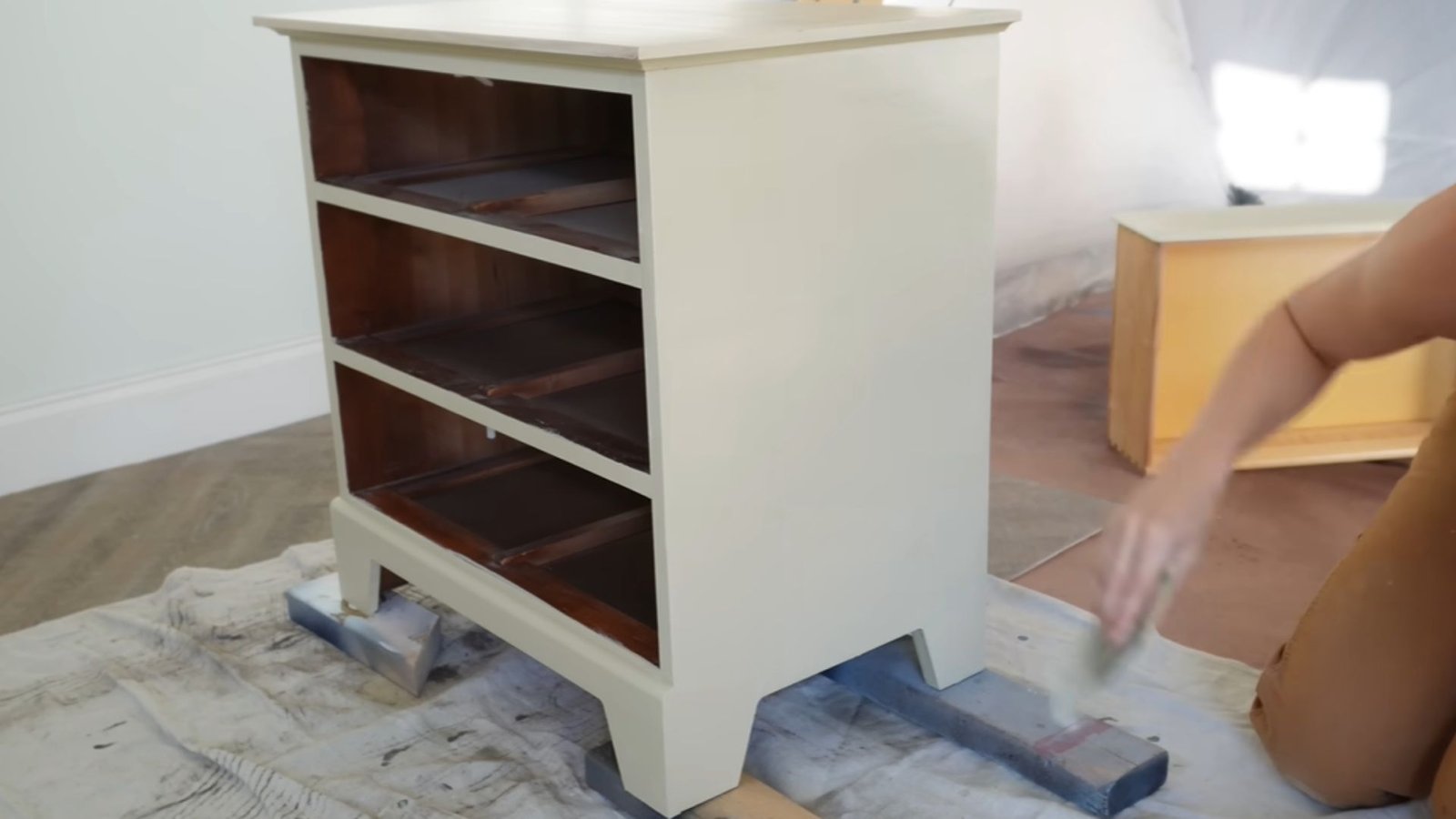 A freshly painted white dresser, without drawers, on a tarp-covered floor. A person is sanding a nearby drawer, suggesting a DIY refurbishing project.